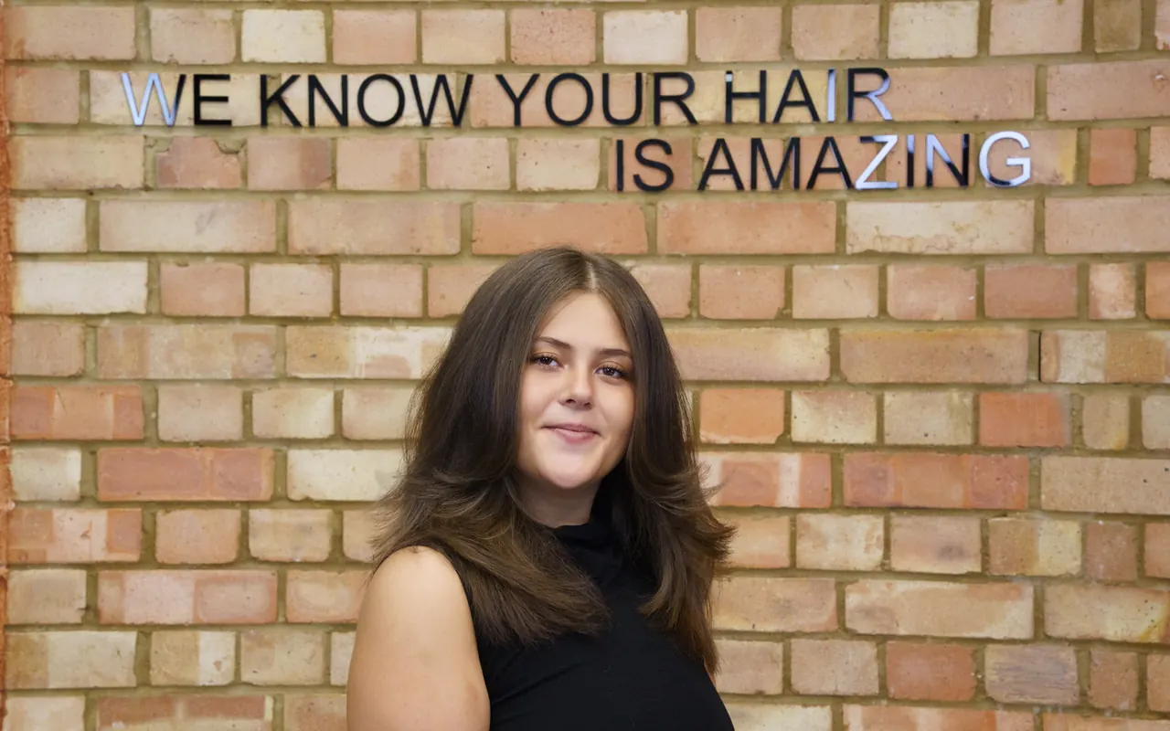 A client with long brown hair under the brick wall that reads “We know your hair is amazing”